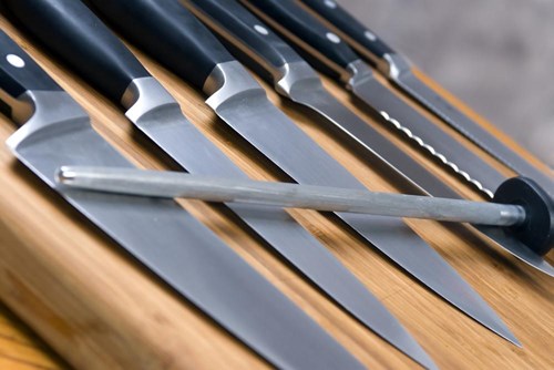 6 knive ligger på et træskærebræt med en knivsliber som ligger på tværs af alle knivene